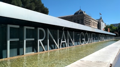Large, bold letters spell out 'Fernán Gómez Centro Cultural' on a modern building facade. A shallow reflective pool stretches alongside the structure. In the background, classical architecture with a dome and a flag are visible. The setting is outdoors, with clear skies and greenery around.