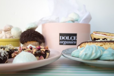An assortment of desserts displayed on plates includes doughnuts with glaze and dried flowers, a cupcake with chocolate frosting, meringue cookies, and a cake slice. A box labeled 'Dolce Organico' holds more treats and is centrally positioned in the background.