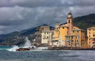 A coastal town featuring colorful buildings with a prominent clock tower, set against a dramatic cloudy sky. The sea is rough, with waves crashing against the rocky shoreline, creating splashes of water. People can be seen walking along the waterfront, enjoying the scenic view.