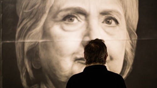A person stands in front of a large grayscale portrait of a woman's face. The focus is on the eyes and the intricate details captured in the artwork. The person's back is to the viewer, and they are observing the portrait closely.