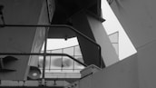 The image presents a black and white industrial scene with various metal beams and structures. Geometric shapes and lines are formed by the intersecting metal components. In the background, a glass building facade with curved reflective surfaces is visible.