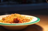 A plate of noodles garnished with green onions and what appears to be a spicy seasoning or paste on one side. The dish is presented on a white plate with a green rim, set against a dimly lit table or countertop.