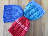 Three knitted hats are arranged on a wooden surface. The hats have a ribbed knitting pattern and come in different colors: one is blue, another is a mixture of blue and green, and the last one is red with orange accents.