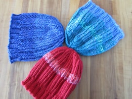 Three knitted hats are arranged on a wooden surface. The hats have a ribbed knitting pattern and come in different colors: one is blue, another is a mixture of blue and green, and the last one is red with orange accents.