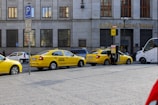 Several yellow taxis are parked along a street near a building with the sign Ceska Narodni Banka. The taxis have 'AAA Radiotaxi' written on their sides and a taxi sign on the roof. A passenger is entering one of the taxis. Next to them, a white bus is partially visible. The area seems to be a designated taxi parking area.