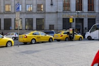 Several yellow taxis are parked along a street near a building with the sign Ceska Narodni Banka. The taxis have 'AAA Radiotaxi' written on their sides and a taxi sign on the roof. A passenger is entering one of the taxis. Next to them, a white bus is partially visible. The area seems to be a designated taxi parking area.