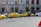 Several yellow taxis are parked along a street near a building with the sign Ceska Narodni Banka. The taxis have 'AAA Radiotaxi' written on their sides and a taxi sign on the roof. A passenger is entering one of the taxis. Next to them, a white bus is partially visible. The area seems to be a designated taxi parking area.