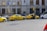 Several yellow taxis are parked along a street near a building with the sign Ceska Narodni Banka. The taxis have 'AAA Radiotaxi' written on their sides and a taxi sign on the roof. A passenger is entering one of the taxis. Next to them, a white bus is partially visible. The area seems to be a designated taxi parking area.