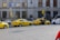 Several yellow taxis are parked along a street near a building with the sign Ceska Narodni Banka. The taxis have 'AAA Radiotaxi' written on their sides and a taxi sign on the roof. A passenger is entering one of the taxis. Next to them, a white bus is partially visible. The area seems to be a designated taxi parking area.