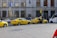 Several yellow taxis are parked along a street near a building with the sign Ceska Narodni Banka. The taxis have 'AAA Radiotaxi' written on their sides and a taxi sign on the roof. A passenger is entering one of the taxis. Next to them, a white bus is partially visible. The area seems to be a designated taxi parking area.