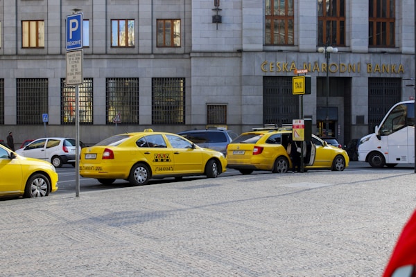 Several yellow taxis are parked along a street near a building with the sign Ceska Narodni Banka. The taxis have 'AAA Radiotaxi' written on their sides and a taxi sign on the roof. A passenger is entering one of the taxis. Next to them, a white bus is partially visible. The area seems to be a designated taxi parking area.
