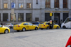 Several yellow taxis are parked along a street near a building with the sign Ceska Narodni Banka. The taxis have 'AAA Radiotaxi' written on their sides and a taxi sign on the roof. A passenger is entering one of the taxis. Next to them, a white bus is partially visible. The area seems to be a designated taxi parking area.