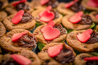 Cupcakes with swirls of chocolate frosting are topped with small red fondant heart decorations. The cupcakes are arranged closely together, creating a colorful and festive presentation.