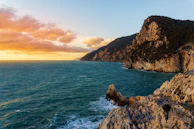 A dramatic shot of rocky cliffs meeting the deep blue sea at sunset.