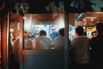 A cozy, dimly-lit ramen shop captures a group of people sitting at a counter. The chef is actively preparing food behind a steaming kitchen, with warm lighting adding an intimate atmosphere. Hanging banners with Japanese characters and various kitchen utensils are visible.