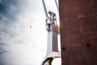A large industrial crane is adjacent to a tall brick wall. The crane is primarily white with a cabin for the operator, and it is positioned against a cloudy sky, suggesting an outdoor setting.