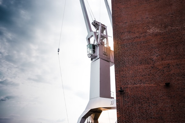 A large industrial crane is adjacent to a tall brick wall. The crane is primarily white with a cabin for the operator, and it is positioned against a cloudy sky, suggesting an outdoor setting.
