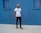 A person stands against a vivid blue wall, wearing a pink cap, white t-shirt with the word 'YOUTH' in colorful letters, black ripped jeans, and sneakers. The setting appears to be urban with a minimalist and stylish vibe.