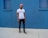 A person stands against a vivid blue wall, wearing a pink cap, white t-shirt with the word 'YOUTH' in colorful letters, black ripped jeans, and sneakers. The setting appears to be urban with a minimalist and stylish vibe.