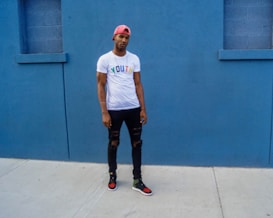 A person stands against a vivid blue wall, wearing a pink cap, white t-shirt with the word 'YOUTH' in colorful letters, black ripped jeans, and sneakers. The setting appears to be urban with a minimalist and stylish vibe.