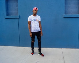 A person stands against a vivid blue wall, wearing a pink cap, white t-shirt with the word 'YOUTH' in colorful letters, black ripped jeans, and sneakers. The setting appears to be urban with a minimalist and stylish vibe.