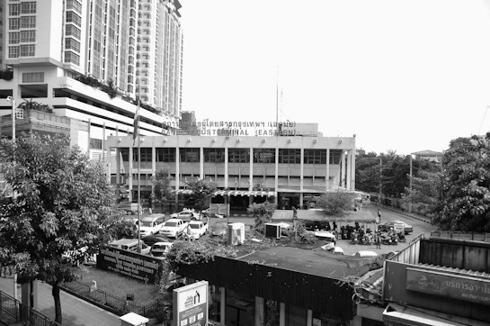 A large building stands prominently in the center, displaying signs in both English and another language, indicating it is a bus terminal. High-rise buildings are visible on the left, with a busy area involving multiple parked cars and motorbikes in the foreground. The area is surrounded by trees and smaller structures, with a clear sky above.