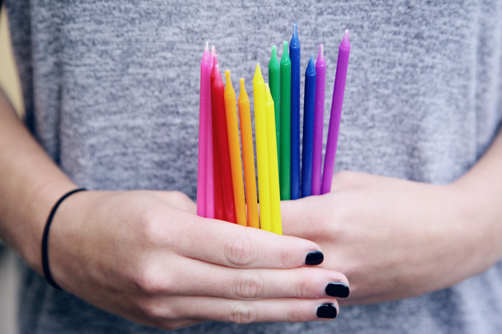 A set of long thin candles are held in clasped hands, ordered by color from pink and red to blue and purple.