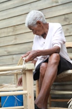 An elderly person is engaged in crafting or repairing a woven chair. The individual appears focused, with hands working on the wooden frame, which is partially bound with weaving material. The background consists of a wooden plank wall giving a rustic feel.