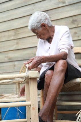 An elderly person is engaged in crafting or repairing a woven chair. The individual appears focused, with hands working on the wooden frame, which is partially bound with weaving material. The background consists of a wooden plank wall giving a rustic feel.