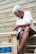 An elderly person is engaged in crafting or repairing a woven chair. The individual appears focused, with hands working on the wooden frame, which is partially bound with weaving material. The background consists of a wooden plank wall giving a rustic feel.