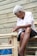 An elderly person is engaged in crafting or repairing a woven chair. The individual appears focused, with hands working on the wooden frame, which is partially bound with weaving material. The background consists of a wooden plank wall giving a rustic feel.