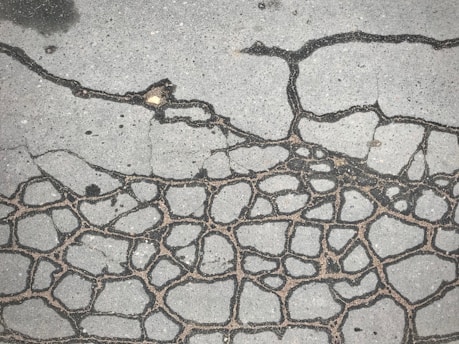 A concrete surface with numerous dark cracks forming intricate patterns across the pavement. The texture is rough, and the cracks appear to outline irregular shapes resembling a network or cells. Slight discolorations and small debris are visible on the surface.