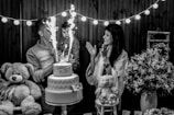 A black and white photo capturing a joyful scene at a celebration, where a man holds a young child near a two-tiered cake with sparklers. A woman stands nearby clapping and smiling. The scene is decorated with strings of lights, large teddy bears, and floral arrangements on the table.