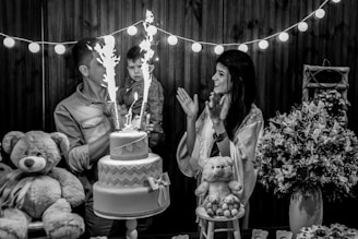 A black and white photo capturing a joyful scene at a celebration, where a man holds a young child near a two-tiered cake with sparklers. A woman stands nearby clapping and smiling. The scene is decorated with strings of lights, large teddy bears, and floral arrangements on the table.