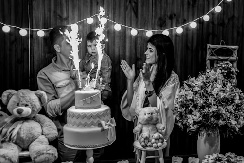 A black and white photo capturing a joyful scene at a celebration, where a man holds a young child near a two-tiered cake with sparklers. A woman stands nearby clapping and smiling. The scene is decorated with strings of lights, large teddy bears, and floral arrangements on the table.