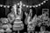 A black and white photo capturing a joyful scene at a celebration, where a man holds a young child near a two-tiered cake with sparklers. A woman stands nearby clapping and smiling. The scene is decorated with strings of lights, large teddy bears, and floral arrangements on the table.