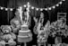 A black and white photo capturing a joyful scene at a celebration, where a man holds a young child near a two-tiered cake with sparklers. A woman stands nearby clapping and smiling. The scene is decorated with strings of lights, large teddy bears, and floral arrangements on the table.