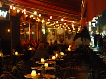 A warmly lit outdoor cafe in the evening, featuring numerous tables with small glowing lanterns. The atmosphere is bustling yet cozy, with strings of colorful lights overhead. The scene includes several people sitting and socializing under a canopy, adding to the lively ambiance.