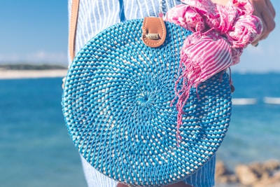 A round, woven blue bag held by a person near a beach. The bag has a leather strap and features a pink and white striped scarf tied to it. The background displays a clear blue ocean with a blurred shoreline.