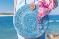 A round, woven blue bag held by a person near a beach. The bag has a leather strap and features a pink and white striped scarf tied to it. The background displays a clear blue ocean with a blurred shoreline.