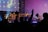 A lively concert scene with a band performing on stage. Bright lights illuminate the musicians, and a large screen displays an image of a guitarist. In the foreground, silhouettes of audience members are visible, with one individual raising their hands in excitement.