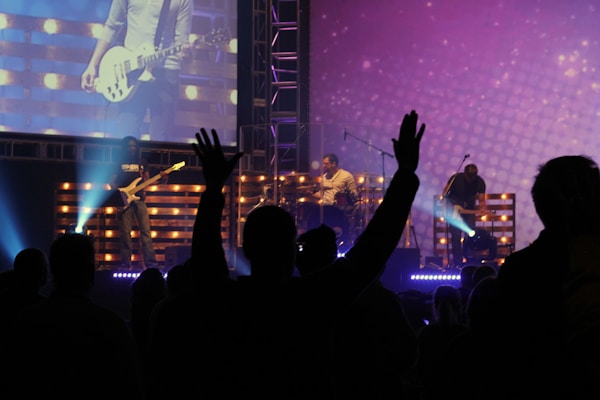 A lively concert scene with a band performing on stage. Bright lights illuminate the musicians, and a large screen displays an image of a guitarist. In the foreground, silhouettes of audience members are visible, with one individual raising their hands in excitement.