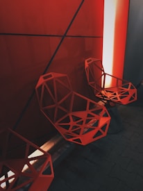 Geometric, angular chairs with a bold, intricate pattern sit against a wall illuminated by a strong red and orange light. The design is modern and striking, featuring an industrial and futuristic aesthetic. The overall color scheme is dominated by shades of red with deep shadows creating an intriguing atmosphere.