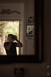 A man is capturing a photo in front of a mirror, with natural light coming from an open door. The background includes a textured wall with photographs and wall decor.