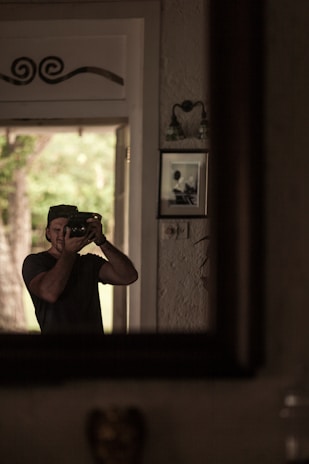 A man is capturing a photo in front of a mirror, with natural light coming from an open door. The background includes a textured wall with photographs and wall decor.