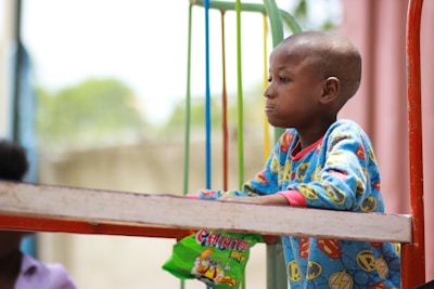 A child wearing a brightly colored outfit with cartoon patterns stands beside a metal rail, holding a green snack package. The background appears soft and blurred, suggesting an outdoor setting.