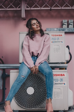 A woman sits comfortably on an air conditioning unit against a pink wall. She wears a pink sweater, ripped blue jeans, and pink slip-on shoes. Her eyes are closed, and she appears relaxed and content.