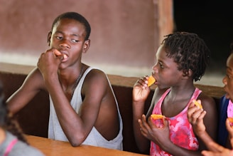 A group of children sitting at a table, eating snacks. The focus is on two children sharing a moment, with one wearing a grey sleeveless top and the other in a patterned pink dress. They appear engaged and contemplative.