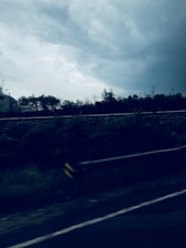 A semi-truck is seen driving along a highway under a cloudy sky. The road is bordered by a guardrail, and there are trees in the background. The atmosphere appears stormy, with dark clouds overhead.
