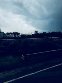 A semi-truck is seen driving along a highway under a cloudy sky. The road is bordered by a guardrail, and there are trees in the background. The atmosphere appears stormy, with dark clouds overhead.